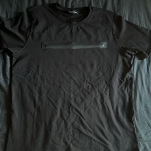 Svart Calvin Klein Jeans t-shirt - Svart t-shirt från Calvin Klein Jeans med stilren logga i svart gummi på bröstet. Klassisk rund hals och korta ärmar. Tillverkad i 100% bomull för skön känsla och bra passform. Perfekt för dig som gillar minimalistisk design och snygga detaljer.