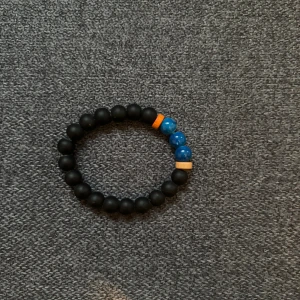 Svart och blått pärlarmband - Stilrent armband med svarta matta pärlor, tre glansiga blå pärlor och två små orange och beige detaljer. Perfekt accessoar för att ge din outfit en cool touch. Elastiskt band gör det enkelt att ta på och av.