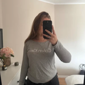 Grå långärmad tröja Calvin Klein - Säljer en stilren grå långärmad tröja från Calvin Klein med vit logotyp framtill. Tröjan har rund hals och är tillverkad i mjuk bomull, perfekt för en chill och enkel look. Passar dig som gillar klassiska och cleana plagg.