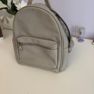 Ljusgrå ryggsäck i skinnimitation - Snygg liten ljusgrå ryggsäck med stilren design och praktisk framficka med dragkedja. Väskan har justerbara axelband.