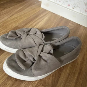 Grå mockasneakers med rosett, strl 38 - Snygga grå sneakers från Alley i mockaimitation med stor rosettdetalj framtill. Skorna har vit platt sula och rund tå, vilket ger en chill och trendig look. Perfekta att slänga på sig till jeans eller kjol för en avslappnad stil. Använda några gånger köpta på skopunkten💕