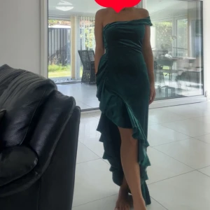 Mörkgrön offshoulder klänning med volang - Säljer en elegant mörkgrön klänning i sammet med offshoulder-design och asymmetrisk volang längs sidan. Klänningen har en lång siluett med slits framtill och sitter snyggt figurnära. Perfekt för dig som vill sticka ut med en unik look.