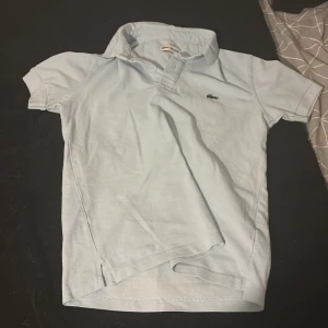 Ljusblå Lacoste pikétröja - Snygg ljusblå pikétröja från Lacoste med klassisk krage och korta ärmar. Tröjan har den ikoniska krokodilloggan broderad på bröstet och är gjord i mjuk bomull. Perfekt för en clean och sportig look.
