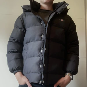 Svart pufferjacka från Gant - Säljer en svart pufferjacka från Gant med huva. Jackan har dragkedja och knappar framtill.  Klassisk och stilren design som funkar till det mesta. Den är knappt använd och har inga defekter. Bra passform 