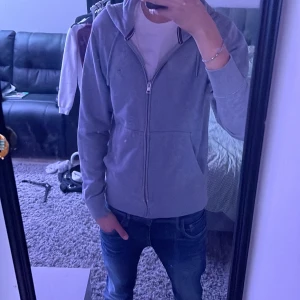 Grå hoodie med dragkedja från Gant - Säljer en stilren grå hoodie från Gant med dragkedja och huva. Tröjan har snörning i huvan, två fickor framtill och en liten broderad Gant-logga på bröstet. Perfekt för en avslappnad look och passar till jeans eller joggers. Det finns ett litet hål vid ärmen men det syns inte vid användning och det är lätt lagat ifall man vill göra det så tveka inte på att höra av er vid minsta lilla fundering👍Pris går att diskuteras vid snabb affär🤩 