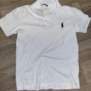 Vit piké från Ralph Lauren - Klassisk vit pikétröja från Ralph Lauren med korta ärmar och svart broderad logga på bröstet. Tröjan har krage och knappar framtill, tillverkad i mjuk bomull. Perfekt för en clean och stilren look.