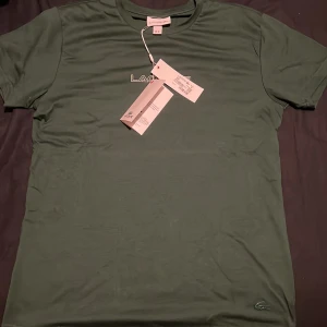Grön Lacoste t-shirt med logga - Snygg grön t-shirt från Lacoste i storlek M. T-shirten har klassisk rund hals, korta ärmar och en stilren vit Lacoste-logga på bröstet. Materialet är mjuk bomull och den ikoniska krokodilen syns nere vid fållen. Perfekt för dig som gillar clean och sportig stil.
