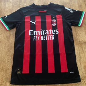 AC Milan matchtröja Puma röd/svart - AC Milan fotbollströja från Puma med klassiska röda och svarta vertikala ränder, klubbmärke broderat på bröstet och vita Puma-loggor. Kortärmad modell i lätt funktionsmaterial med italienska flaggdetaljer på ärmsluten och sponsortryck på bröstet. Buda gärna, pris kan sänkas vid snabb affär!
