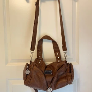 Brun axelväska  - Snygg brun axelväska från Ouyalu i skinnimitation med flera fack och dragkedjor. Väskan har både handtag och justerbar axelrem, samt snygga metalldetaljer och logga framtill. Perfekt storlek för allt du behöver ha med dig och passar till många olika outfits. Jag har bara använt väskan en eller två gånger. 