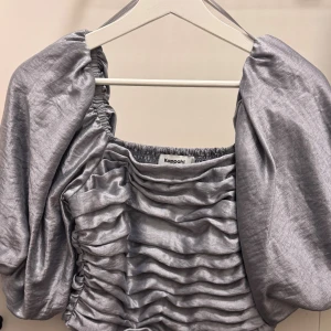 Silvrig puffärmad topp från Kappahl XS - Supersnygg silvrig topp från Kappahl i storlek XS. Modellen har puffiga korta ärmar, smockad rygg och framsida med rynkade detaljer. Toppens material har en glansig finish som ger en lyxig känsla och den fyrkantiga ringningen gör den extra trendig.