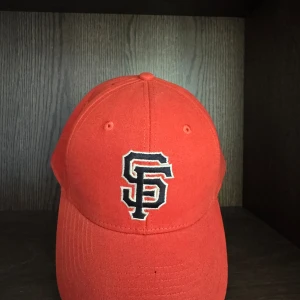  San Francisco Giants keps - Snygg röd keps med broderad SF-logga i svart och vitt framtill. Klassisk böjd skärm och ventilerande hål upptill. Perfekt för dig som vill ha en sportig och stilren look. Tillverkad i bomull för skön passform.