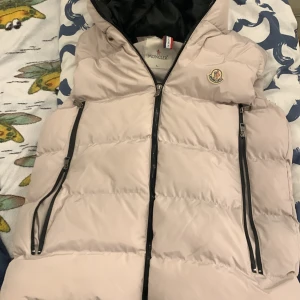 Beige pufferjacka från Moncler L - Snygg beige pufferjacka från Moncler med svart innerfoder och dragkedjor. Jackan har huva, två stora fickor med dragkedja och Moncler-logga på bröstet. Insidan har coolt Moncler-tryck och etiketter. Perfekt för kalla dagar och riktigt stilren look. Vi kan diskutera pris under sms