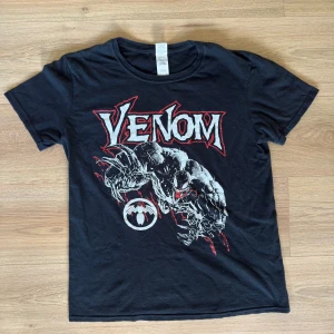 Svart Venom t-shirt från Gildan - Cool svart t-shirt från Gildan med Venom-tryck i vitt och rött på framsidan. Klassisk rund hals och korta ärmar. Materialet är mjuk bomull och passformen är normal. Perfekt för dig som gillar Marvel och vill sticka ut med en snygg graphic tee.