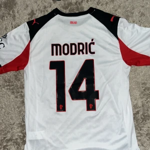 AC Milan Modric 14 bortatröja Puma L - AC Milan bortatröja från Puma med Modric och nummer 14 på ryggen. Tröjan är vit med svarta och röda detaljer, svart krage och korta ärmar. Tillverkad i lätt och ventilerande DryCell-material, perfekt för match eller träning. Snyggt tryck med Emirates Fly Better på bröstet.