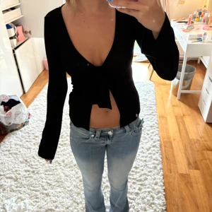 Svart omlottopp med knyt framtill - Svart långärmad omlottopp med djup v-ringning och knyt framtill. Toppen är croppad och har en tight passform som framhäver midjan. Perfekt för dig som gillar en stilren och trendig look. Materialet känns mjukt och stretchigt, troligen bomull eller polyester. Den är i storlek xs och från Gina tricot. Den är lite nopprig kring bysten och armhålorna.