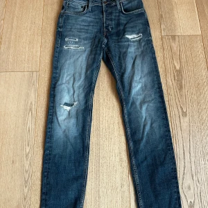Jack & Jones blå tapered jeans 28/32 - Snygga blå jeans från Jack & Jones i modellen Tapered/Mike. Jeansen har slitna detaljer och en tvättad look, med klassisk femficksdesign och normalhög midja. Perfekta för dig som gillar en avslappnad men ändå stilren vibe.