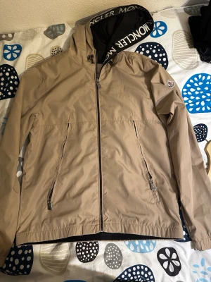 Beige Moncler vindjacka med huva - Stilren beige vindjacka från Moncler med dragkedja framtill, två snedställda fickor med dragkedja och justerbar huva med Moncler-logga. Lätt material som passar perfekt för blåsiga dagar. Klassisk Moncler-patch på ärmen och snygga detaljer på dragkedjorna.