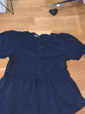 Mörkblå blus med knyt fram - Supersöt mörkblå blus med korta puffärmar och flera knyt framtill. Blusen har en lös passform och lätt rynkad nederdel som ger en flowy känsla. Perfekt att styla med jeans eller kjol för en trendig look.