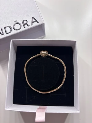 Pandora armband  - Snyggt guldfärgat armband från Pandora med klassisk ormkedja och hjärtformad låsdetalj. Perfekt för att kombinera med berlocker och skapa en personlig stil. Levereras i ask.