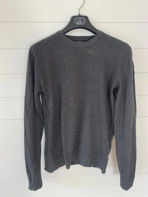 Mörkgrå basic tröja i bomull - En stilren mörkgrå tröja med rund halsringning och långa ärmar. Tröjan är tillverkad i mjuk bomull och har ribbade muddar vid ärmslut och nederkant. Perfekt för lager-på-lager och enkel att matcha med olika outfits.