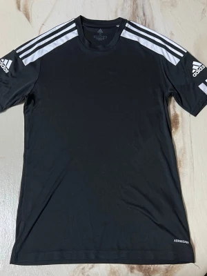 Svart Adidas sporttröja S - Svart sporttröja från Adidas med vita ränder på axlarna och logga på båda ärmarna. Tillverkad i återvunnet Primegreen-material och har Aeroready-teknologi som håller dig torr under träningen. Perfekt för fotboll eller gym.