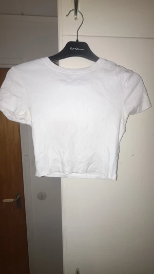 Vit croppad ribbad topp - Säljer en enkel och stilren vit croppad topp med korta ärmar. Toppen är ribbad och har en rund halsringning. Ganska använd och de syns att lite färg gått bort vid texten.