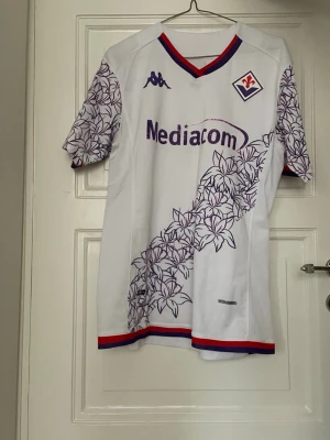 Fiorentina matchtröja - Snygg vit Fiorentina matchtröja från Kappa med lila blommönster på ärmar och snett över framsidan. Röd och lila kant vid hals och ärmslut, samt klubbmärke och sponsortryck på bröstet. Tillverkad i lätt och ventilerande material, perfekt för fotboll eller träning.