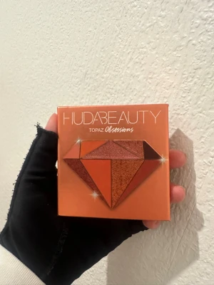 Huda Beauty palett ny - Säljer en Huda Beauty Topaz Obsessions ögonskuggspalett med nio nyanser i varma toner som guld, koppar, brons, orange och brunt. Paletten innehåller både matta och skimrande skuggor i ett kompakt format. Paletten är inte använd. 