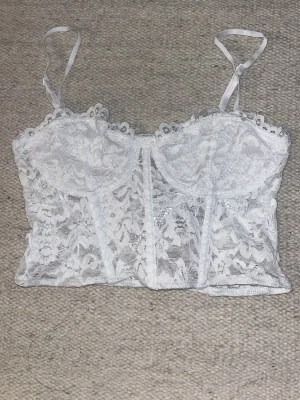 Vit spets corset - Säljer en vit corset i genomskinlig spets med blommigt mönster och tunna justerbara axelband. Corseten har bygel under bysten för extra stöd och en romantisk look. Perfekt för dig som gillar feminina detaljer och snygg passform.