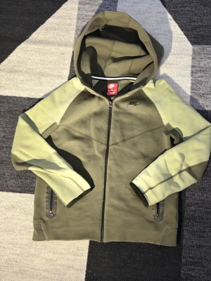Olivgrön hoodie från Nike med dragkedja - Snygg olivgrön hoodie från Nike med svarta detaljer och dragkedja framtill. Hoodien har två fickor med dragkedja och en klassisk huva. Tillverkad i mjukt material som passar perfekt till en avslappnad streetstyle. Storlek 158-170