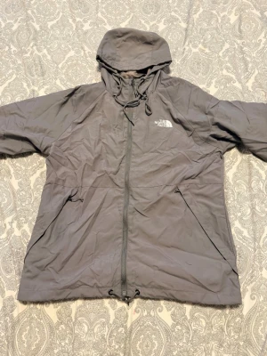 Grå regnjacka från The North Face - Snygg grå regnjacka från The North Face med huva och hel dragkedja framtill. Jackan har två sidofickor med dragkedja och justerbar dragsko i midjan. Tillverkad i ett lätt och vattenavvisande material, perfekt för regniga dagar.