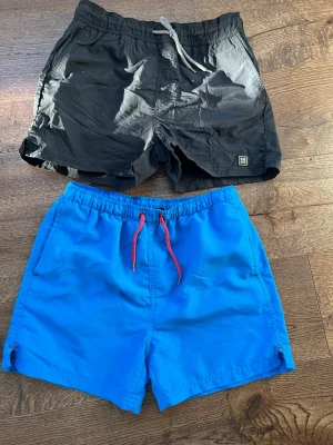 2-pack badbyxor från Lindex, svart & blå - Säljer två par badbyxor från Lindex i storlek 170. Ena paret är helsvart med vitmönstrad dragsko och meshfoder, det andra är klarrött med blått tyg och röd dragsko. Den svarta st 170, den blåa 160