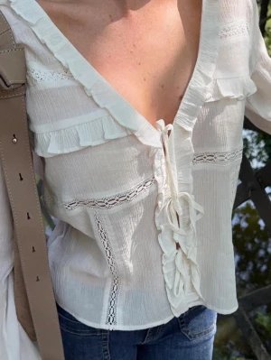 Långärmad vit topp / blus - Säljer en jättefin top/blus från Nelly i storlek M. Blusen är helt oanvänd, endast testad, men lappen är tyvärr borttagen. Den har aldrig kommit till användning.   Väldigt fin modell med volanger längs ringningen, knytning framtill, fina spetsdetaljer, lätt transparens och luftig kvalitet och vida ärmar.   Perfekt till jeans för vardag eller till sommaren, fest och semester.