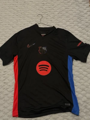 FC Barcelona svart matchtröja Nike - Snygg svart FC Barcelona matchtröja från Nike med röda och blå sidopaneler. Tröjan har klubbmärke och Nike-logga på bröstet samt Spotify-logga framtill. Tillverkad i polyester, perfekt för fotbollsträning eller match.
