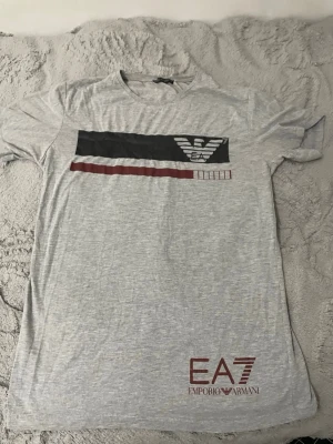 Grå EA7 Emporio Armani t-shirt - Snygg ljusgrå t-shirt från EA7 Emporio Armani med tryck i svart, vitt och rött på bröstet samt logga nere vid fållen. Klassisk rund hals och korta ärmar. Perfekt för dig som gillar stilrena märkesplagg.