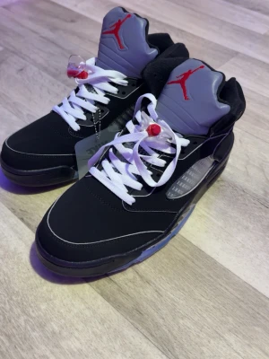 Air Jordan 5 Retro Black/Red - Snygga Air Jordan 5 Retro sneakers i svart med grå och röda detaljer. Skorna har vit sula med isblå genomskinlig undersida, vit snörning och ikonisk Jumpman-logga på plösen. Materialet är en mix av läder och syntet, perfekt för dig som gillar streetwear.