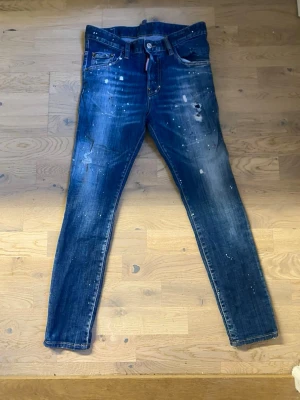 DSQUARED2 blå slim fit jeans - Snygga blå slim fit jeans från DSQUARED2 med slitningar, färgstänk och en cool tvätt. Klassisk femficksmodell med knappgylf och röd DSQ2-lapp bak i midjan. Perfekta för dig som gillar streetstyle och unika detaljer.
