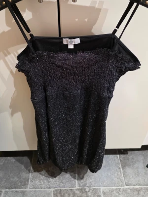 Glittrig svart kort klänning H&M - Snygg svart kort klänning från H&M med glittrigt tyg och smock upptill. Klänningen har smala axelband och en figurnära passform som ger en trendig look. Perfekt för dig som vill sticka ut med lite extra glitter.