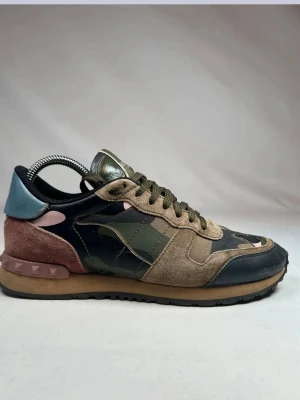 Valentino Garavani camo sneakers - Snygga sneakers från Valentino Garavani med camomönster i grönt, beige och svart. Skorna har detaljer i mocka, läder och textil samt rosa och blå inslag vid hälen. Gummisula med nitar bak ger en unik touch. Perfekta för dig som vill sticka ut med exklusiva sneakers. Pris kan diskuteras