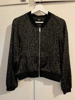 Leopardmönstrad bomberjacka Bik Bok - Snygg leopardmönstrad bomberjacka från Bik Bok i svart och mörkgrått. Jackan har dragkedja framtill, ribbade muddar vid ärmslut och nederkant samt en klassisk bomberkrage. Materialet är tunt och mjukt, perfekt för en trendig streetstyle-look.