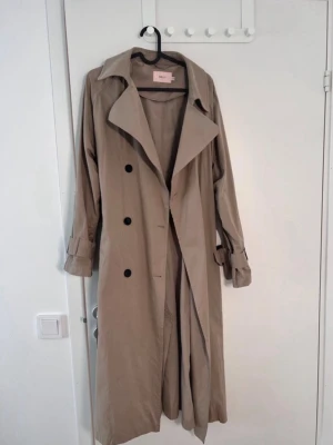 Beige trenchcoat från nelly - Stilren beige trenchcoat från nelly med klassisk krage, knappar framtill och bälte i midjan. Lång modell med långa ärmar och justerbara ärmslut. Perfekt för lager-på-lager och snygg till alla outfits.