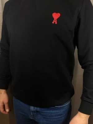 Svart stickad tröja från Ami Paris - Svart stickad tröja från Ami Paris med röd broderad logga i form av ett hjärta och bokstaven A på bröstet. Tröjan har rund halsringning och långa ärmar. Perfekt för en stilren och trendig look.