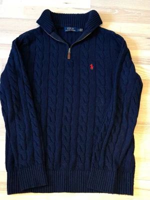 Mörkblå kabelstickad tröja Polo Ralph Lauren quarter zip - Det står storlek L men jag skulle säga att den passar M bättre, är själv 180cm och den var lite stor, men kan funka för L beroende på hur man gillar att det sitter. Kabelstickad mörkblå tröja från Polo Ralph Lauren med halv dragkedja och hög krage. Tröjan har röd broderad logga på bröstet och ribbade muddar vid ärmslut och nederkant. Perfekt för lager-på-lager och klassisk stil.