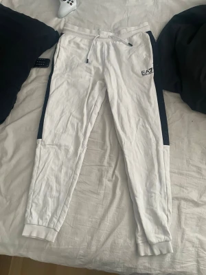 Vita EA7 mjukisbyxor med svarta detaljer - Snygga vita mjukisbyxor från EA7 med svarta paneler längs sidorna och dragsko i midjan. Byxorna har smal passform, muddar vid benslut och EA7-logga på vänster lår. Perfekta för en chill och sportig stil. Har en grön fläck på baksidan, orkar inte tvätta därför vill ha iväg den snabbt. Hör av dig vid intresse 