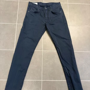 Mörkblå chinos från J.Lindeberg - Snygga mörkblå chinos från J.Lindeberg med klassisk femficksdesign och diskret logga på myntfickan. Byxorna har rak passform och är tillverkade i ett mjukt bomullsmaterial med lite stretch för extra komfort. 31/32