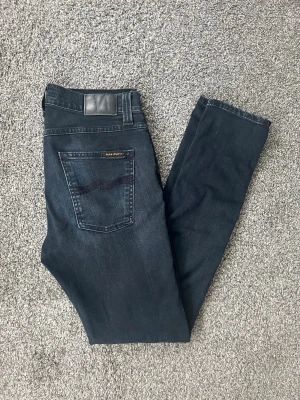 Mörkblå jeans från Nudie Jeans - Säljer ett par mörkblå jeans från Nudie Jeans med klassisk femficksmodell och subtila slitningar framtill. Jeansen har en snygg tvätt och är tillverkade i stretchig denim för extra komfort. Perfekta för dig som gillar en smal passform och stilren look.