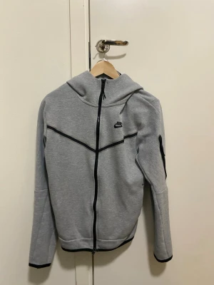 Grå Nike hoodie med dragkedja - Snygg grå hoodie från Nike med svart dragkedja och svarta detaljer. Hoodien har huva, diskret Nike-logga på bröstet och en cool svart rand över bröstet. Tillverkad i mjukt sweatshirtmaterial och har ficka med dragkedja på ärmen. Priset an diskuteras! 