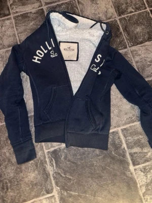 Hollister zip up med ull - Mjuk och varm Hollister zip-hoodie i mörkblå färg. Fodrad med teddyfleece på insidan – perfekt för kyliga dagar. Snygg broderad logga framtill.  Info: • Märke: Hollister • Storlek: S • Färg: Mörkblå • Modell: Zip-hoodie med huva • Skick: Bra begagnat skick