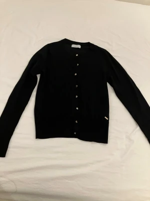 Svart kofta med knappar storlek xs - Stilren svart kofta med rund halsringning och knäppning framtill. Långärmad modell i 77% lyocell 23% polyester, perfekt att slänga över en t-shirt eller linne. Enkel och klassisk design som funkar till det mesta.