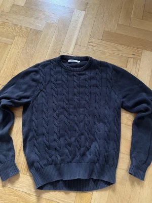 Stickad tröja från dressmann - Mörkblå stickad tröja med klassiskt kabelstickat mönster framtill. Tröjan har rund halsringning, långa ärmar och ribbade muddar vid ärmslut och nederkant. Perfekt för lager-på-lager och en stilren look. Står storlek M men passar även L. Mått längd: 59 cm bredd: 49 cm 